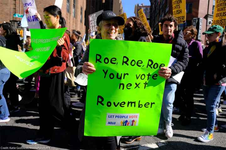 A woman holding a sign reading "Roe, Roe, Roe, Your vote next Rovember / LET PEOPLE VOTE ACLU" poses for the camera as fellow activists walk behind her.