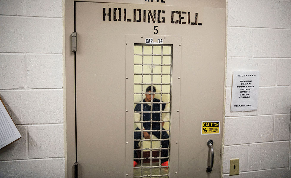 Man in prison cell with door closed, "holding cell" written on the door